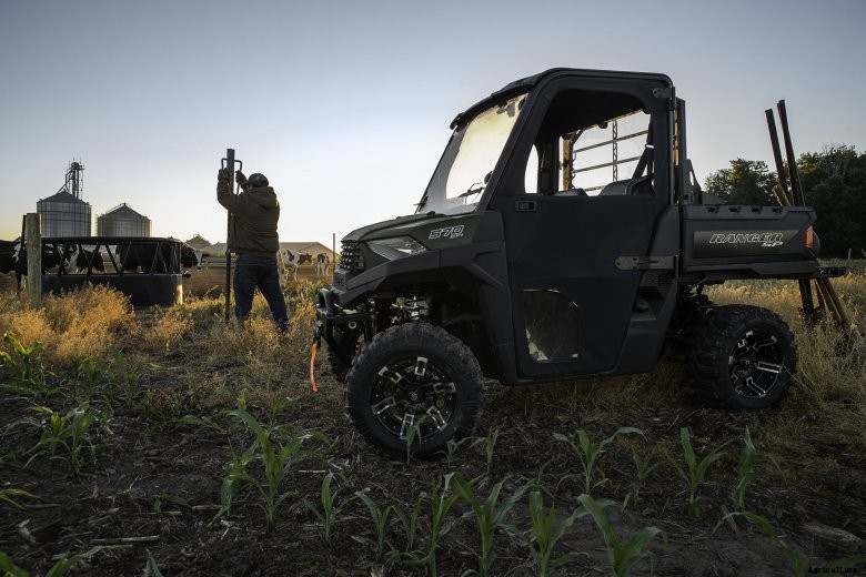 A farmer pounds in fence posts with a Polaris Ranger SP 570 with black cattle in the background